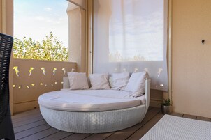 Una cama redonda y blanca de mimbre con muchas almohadas sobre una terraza de madera, cerca de una ventana con vistas a árboles.