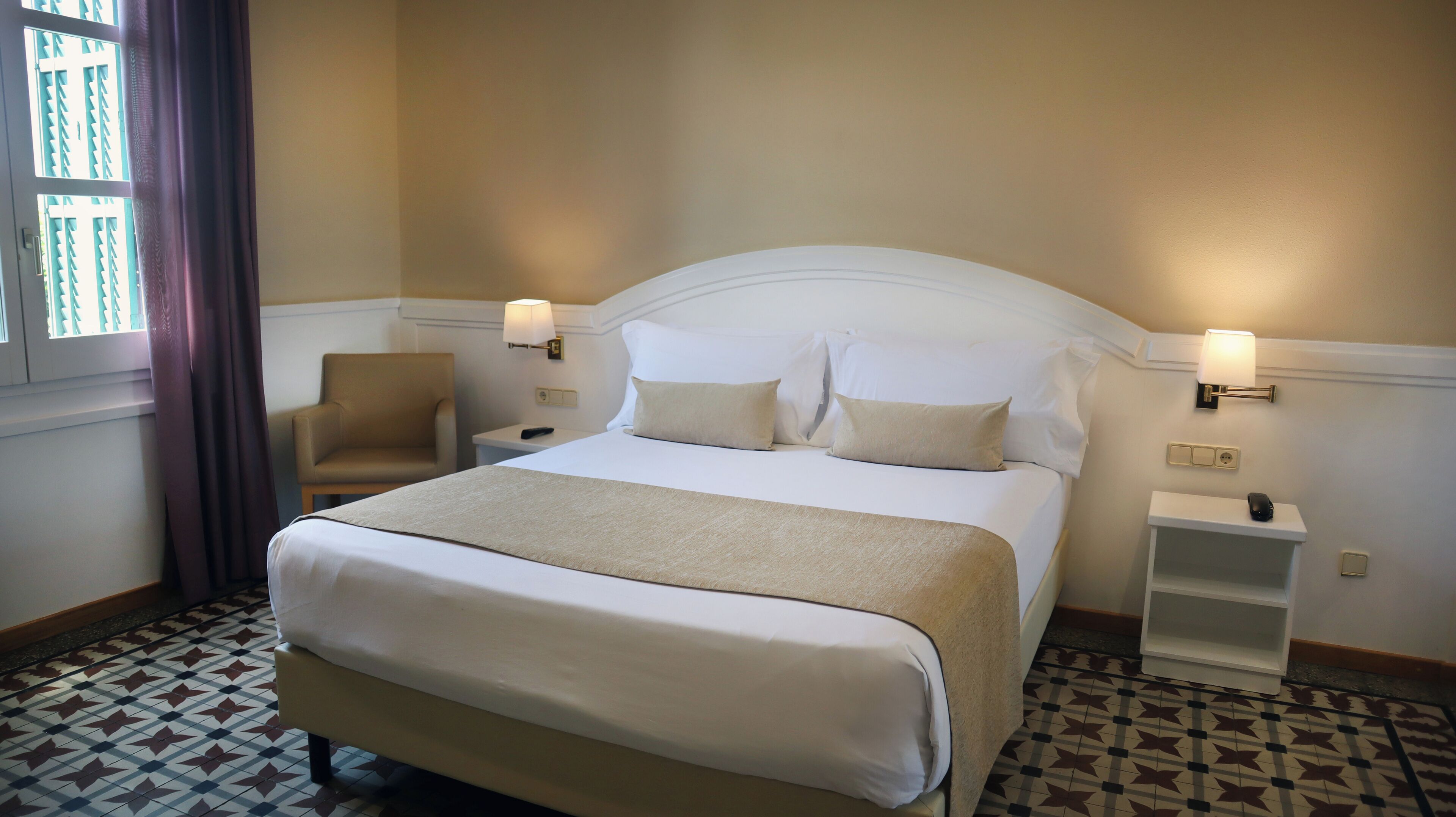 Een hotelkamer met een groot bed, twee nachtkastjes, een stoel en een raam met gordijnen.
