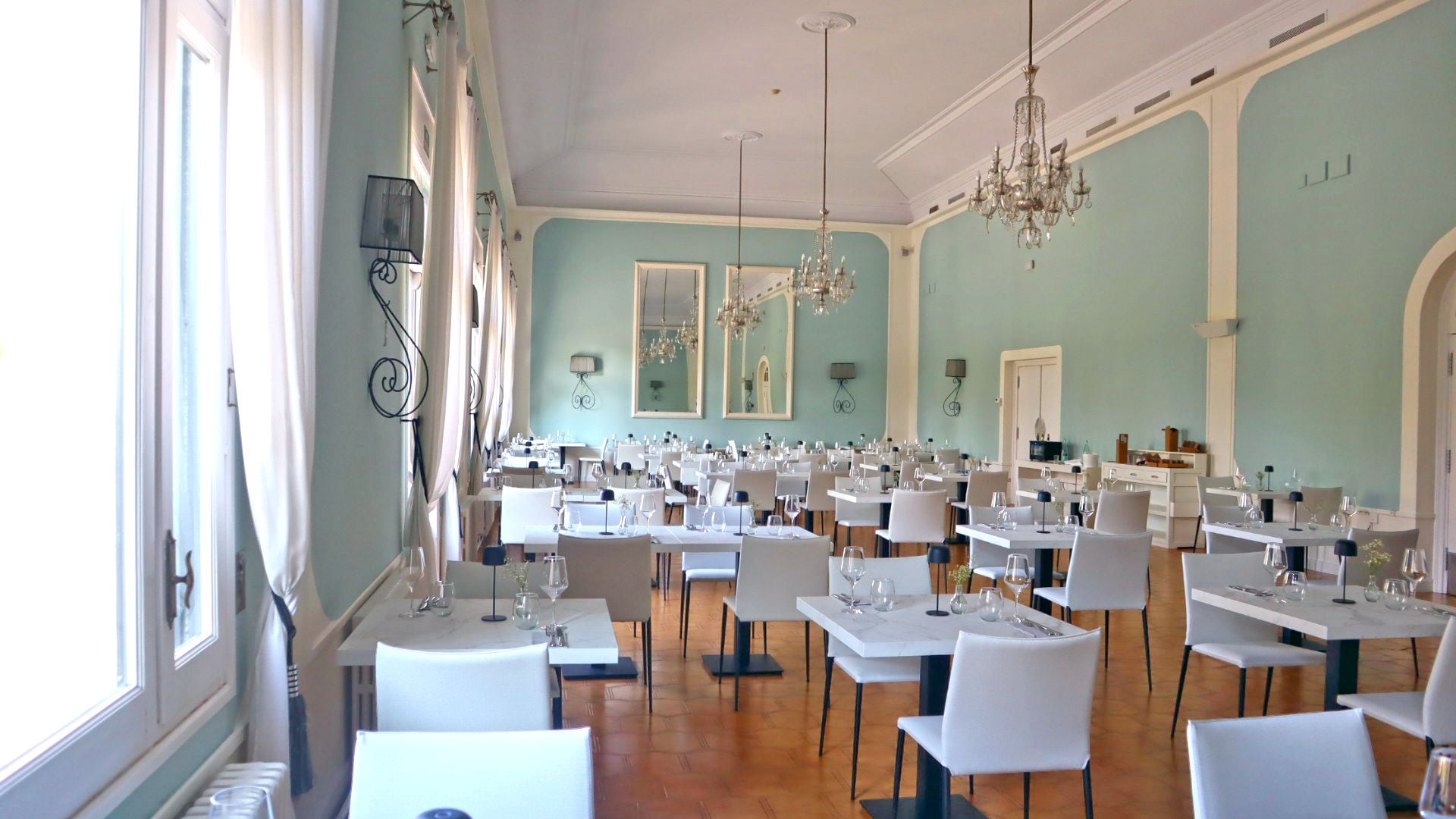 A spacious dining area with multiple tables and chairs, large windows, and elegant chandeliers.
