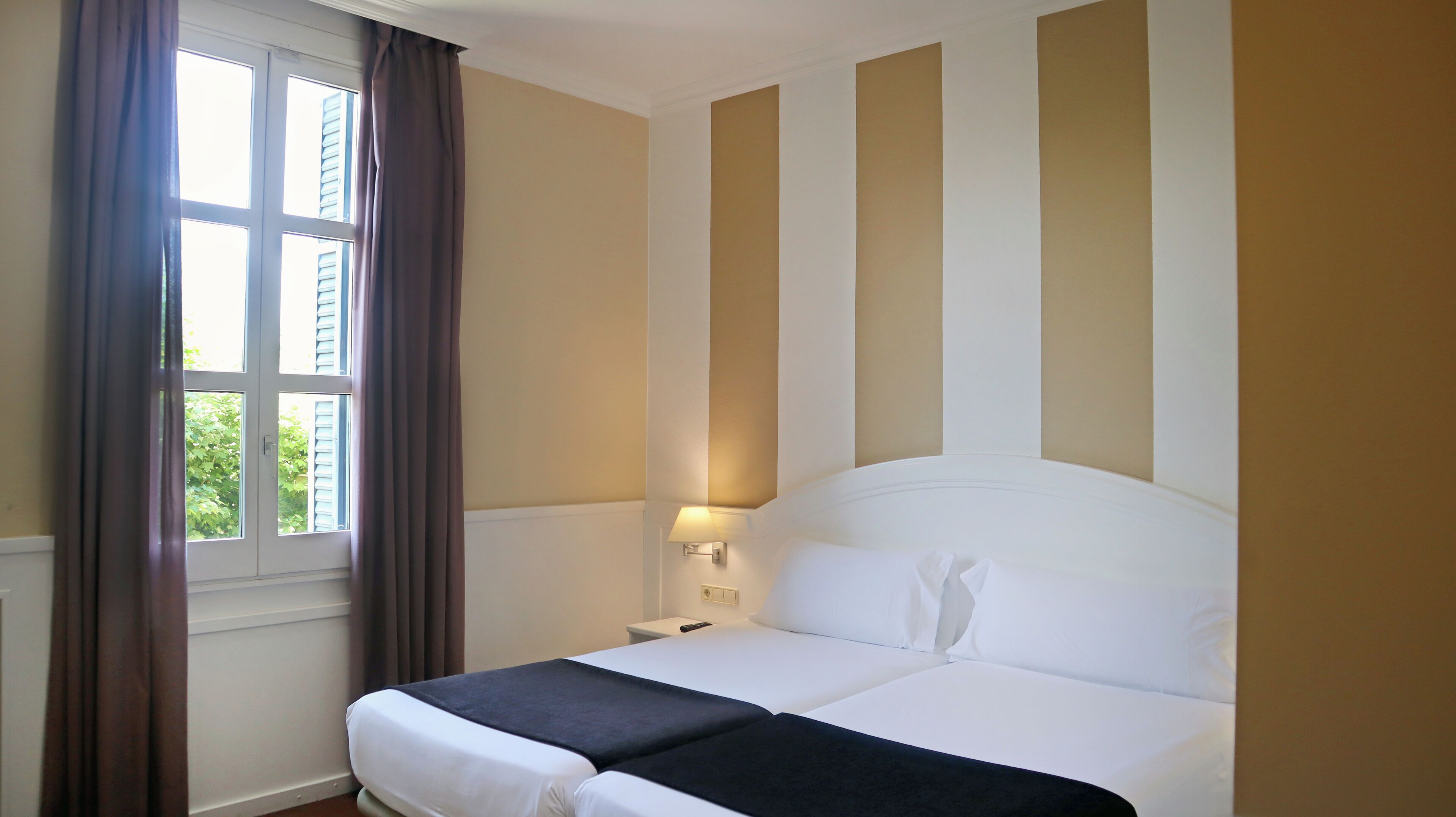 A hotel room with a bed, a nightstand, a lamp, and a window with curtains.
