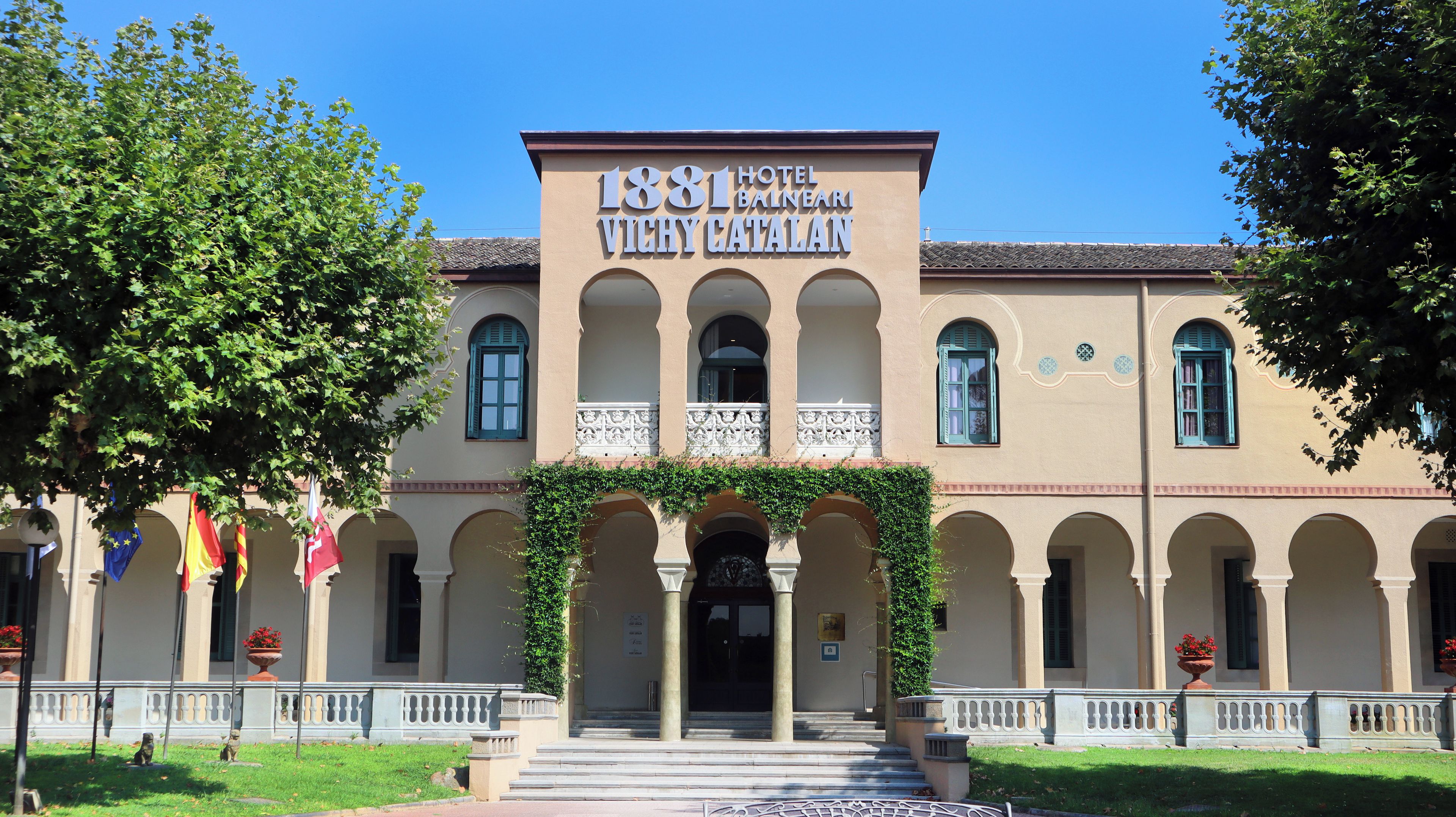 Um edifício histórico com uma placa indicando '1881 Hotel Balneari Vichy Catalan'. Possui uma entrada central com arcos, uma varanda com guarda-corpos decorativos e um telhado em azulejos.