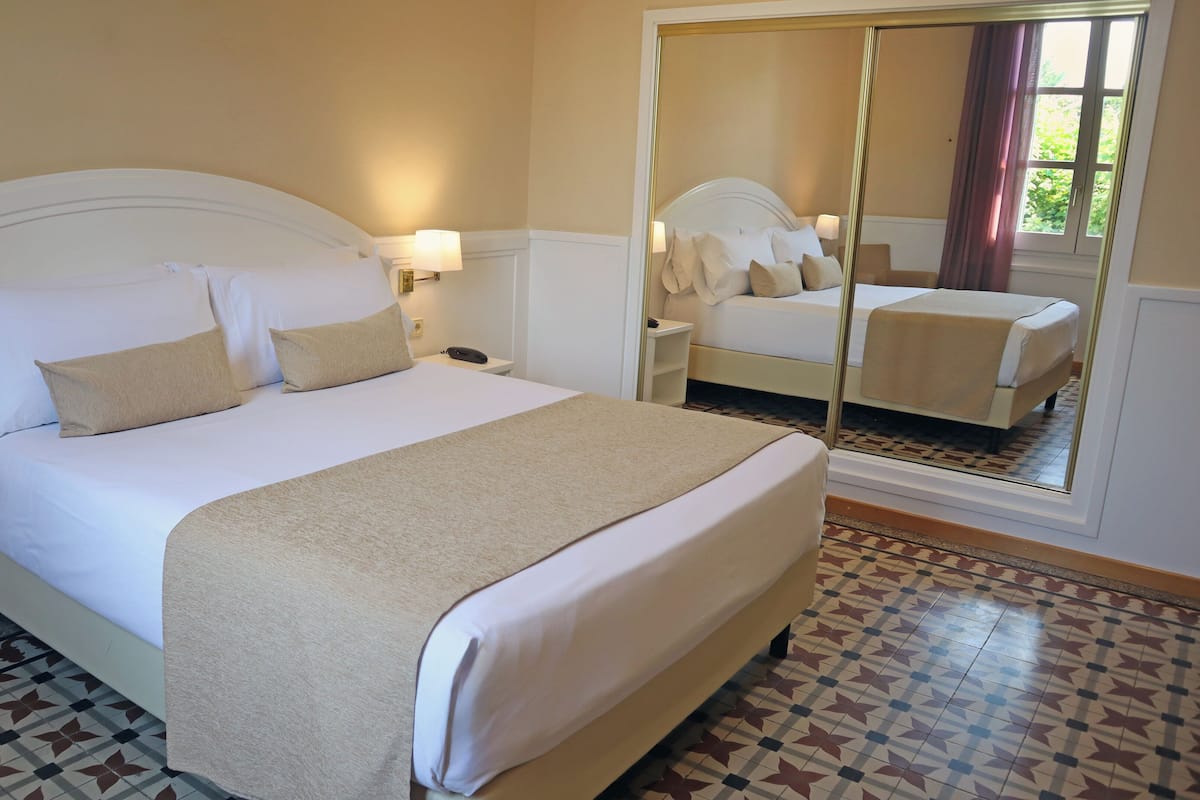 Uma cama bem arrumada com cabeceira branca, mesa de cabeceira com abajur e um espelho grande refletindo o quarto.