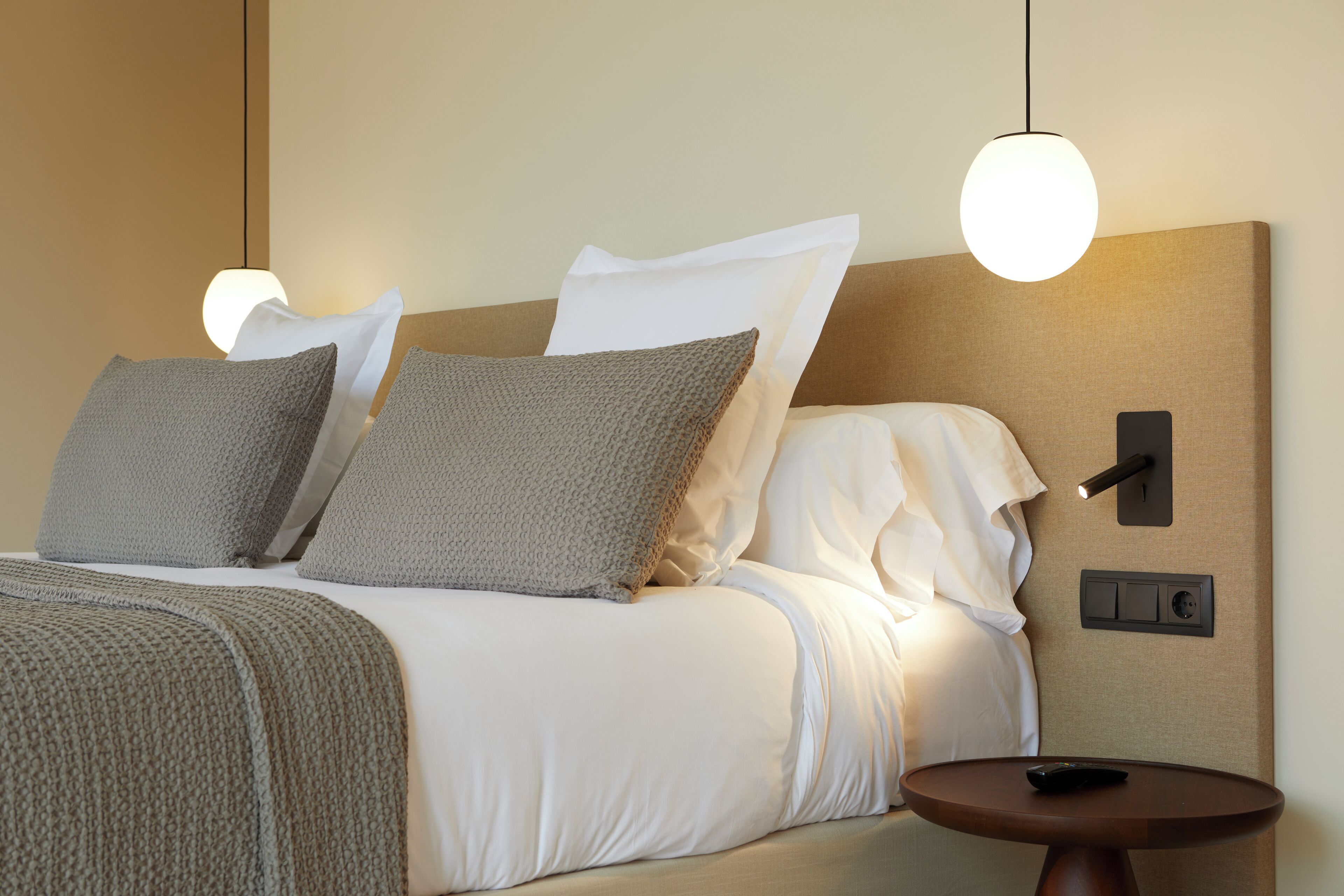 Una cama bien hecha con ropa de cama blanca y gris, una mesita de noche redonda de madera y una lámpara de pared.