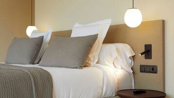 Una cama bien hecha con ropa de cama blanca y gris, una mesita de noche redonda de madera y una lámpara de pared.