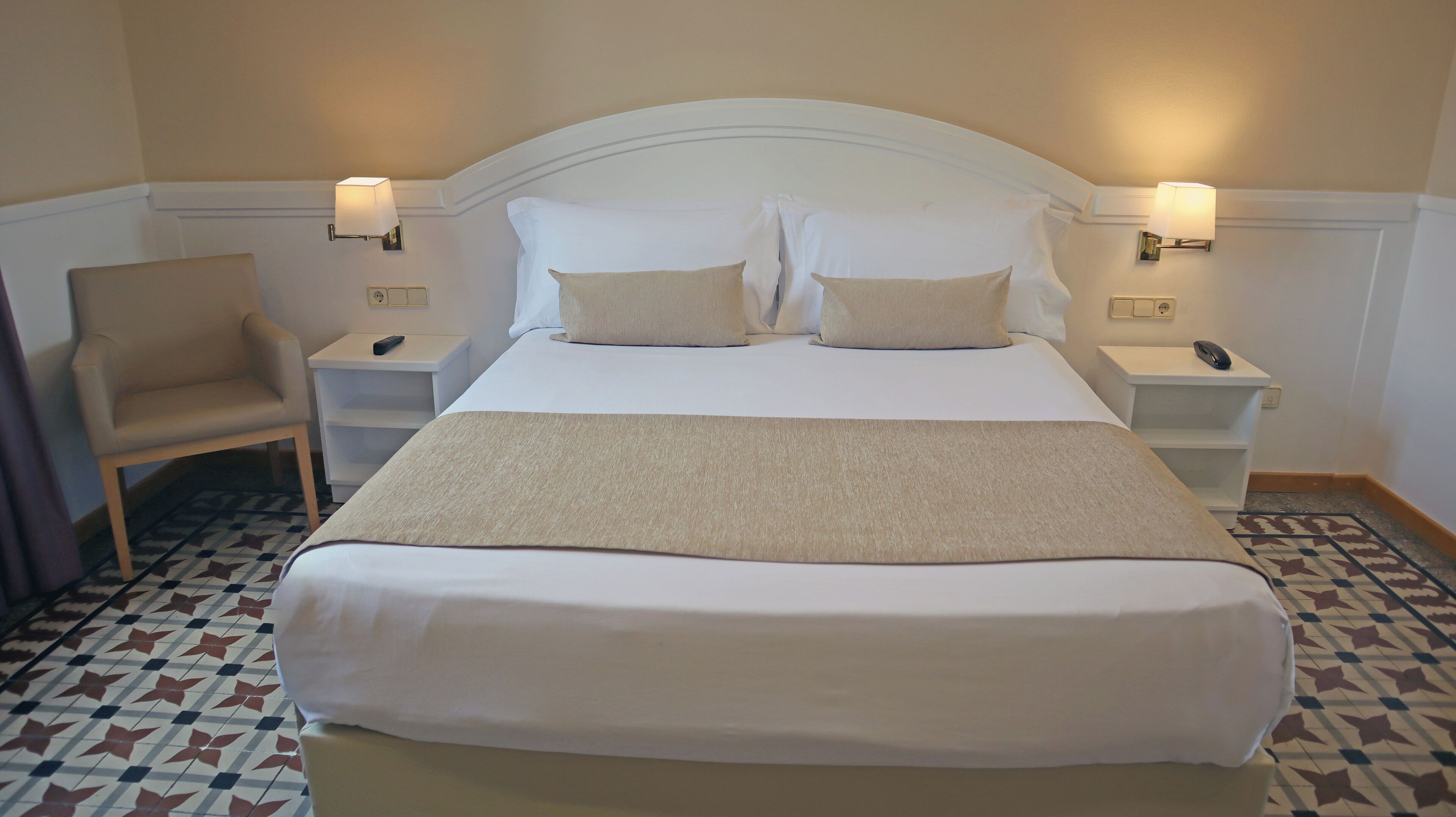 A neatly made bed with a white headboard, two bedside tables with lamps, a chair, and a patterned floor.