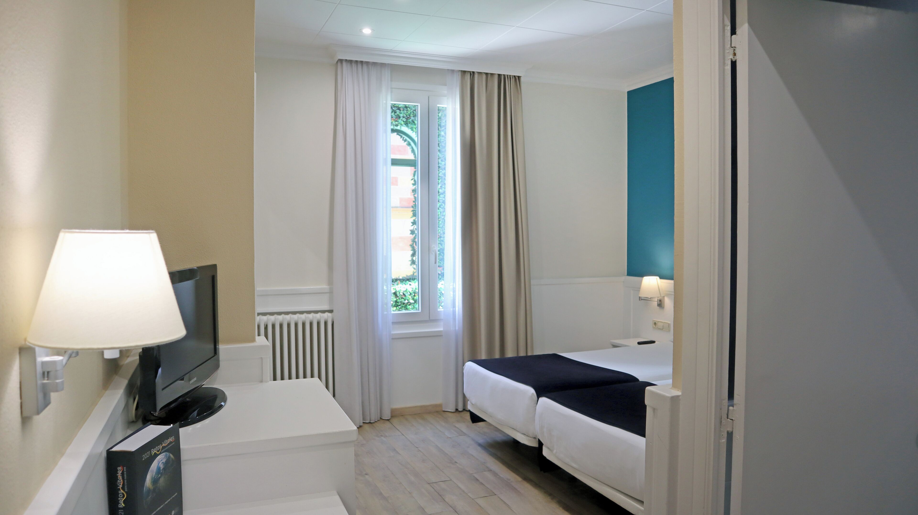 A hotel room with a bed, a desk with a lamp, a TV, and a window with curtains.