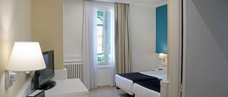 A hotel room with a bed, a desk with a lamp, a TV, and a window with curtains.