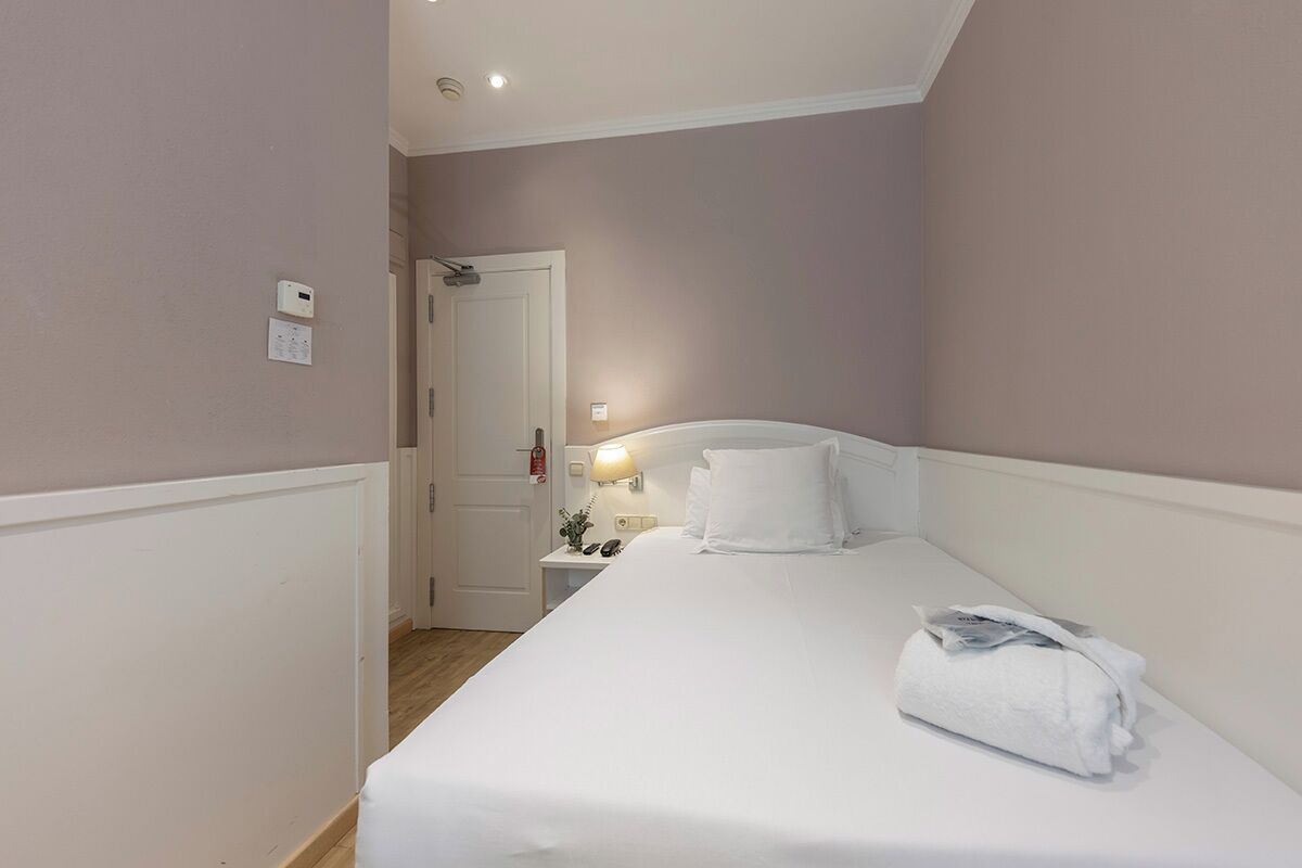 Un dormitorio pequeño y ordenado con una cama individual, una mesita de noche con lámpara, y una puerta que da a otra habitación.