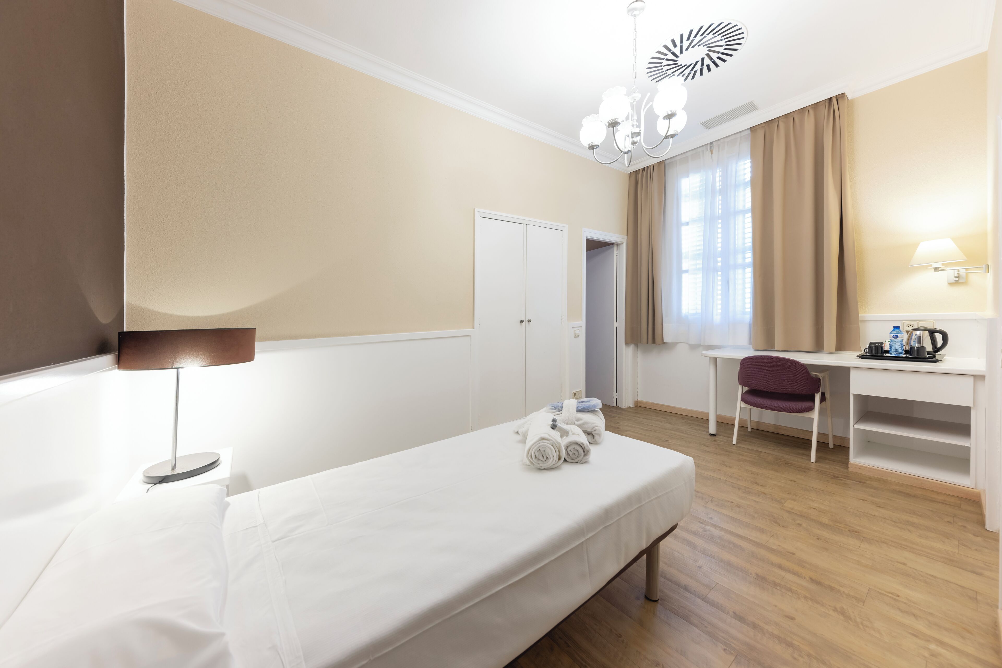 Et hotelværelse med seng, skrivebord, stol, lampe og et vindue med gardiner.