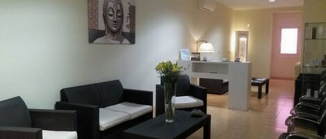 Un área de recepción con mostrador, una sala de espera con sillas, un sofá, una mesa de centro y un jarrón con flores.