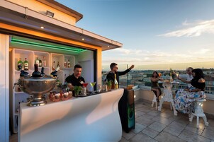 A rooftop bar with a bartender, a bar counter with various bottles and glasses, and guests seated at tables enjoying drinks.