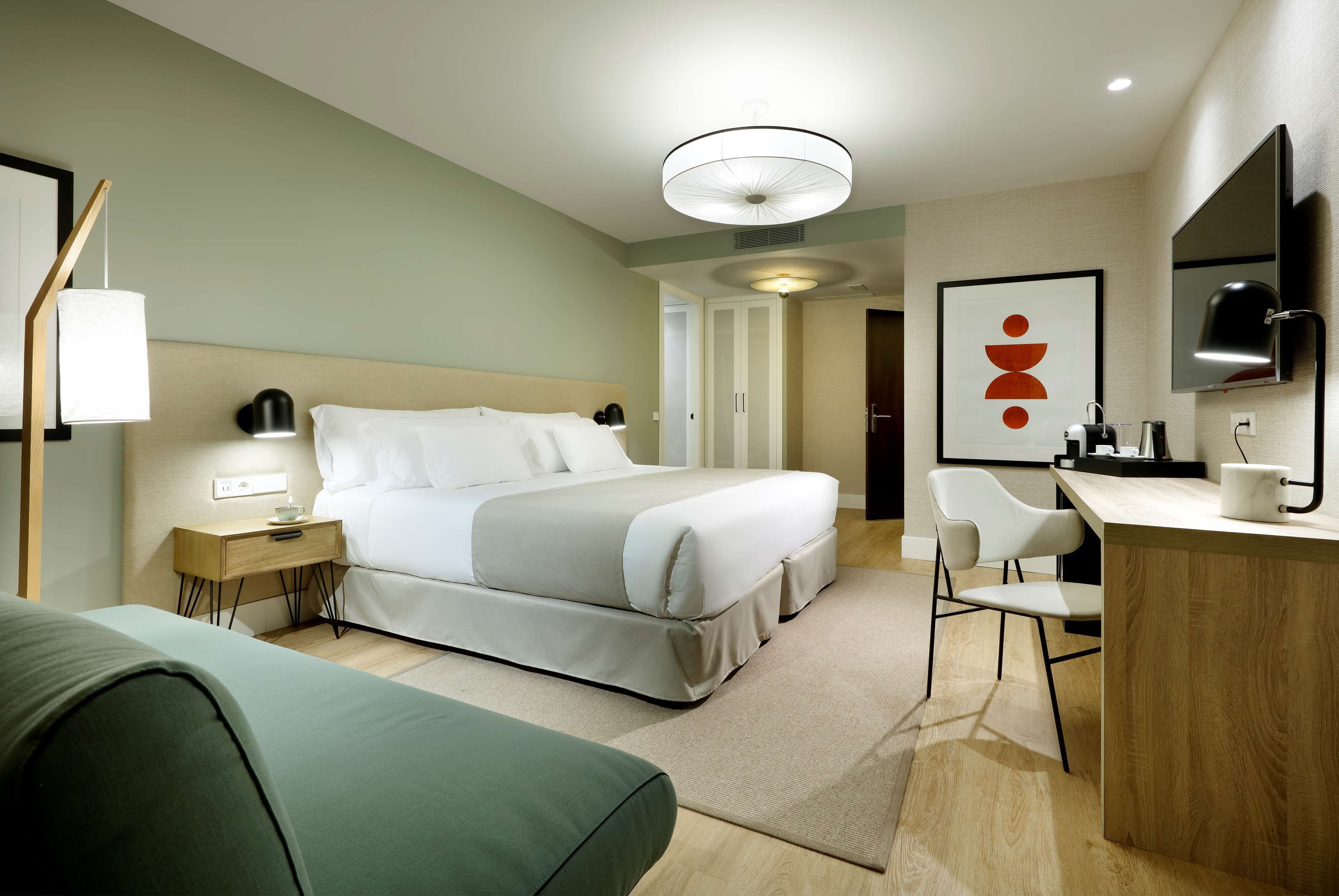 Una habitación de hotel moderna con una cama grande, un escritorio y una silla. Hay una mesita de noche con una lámpara y un televisor montado en la pared.