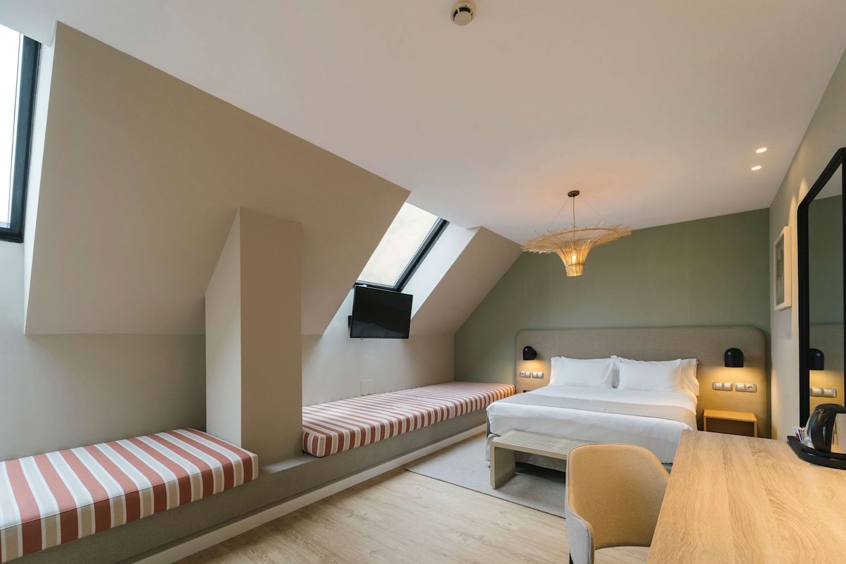 Une chambre moderne avec un plafond en pente, un grand lit, un banc avec des coussins à rayures, une table en bois et un téléviseur fixé au mur.