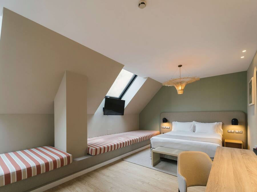 A modern hotel room with a sloped ceiling, a large bed, a bench, a desk, and a TV mounted on the wall.