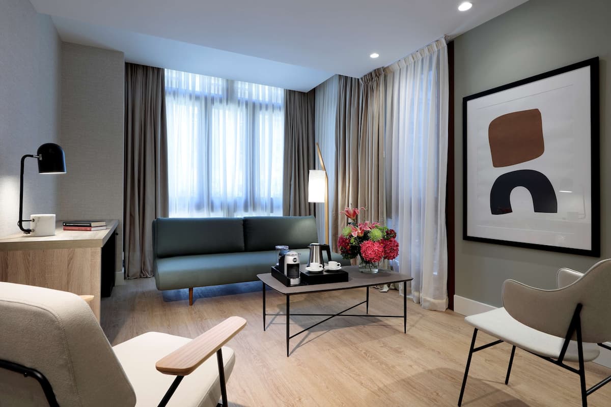 Een moderne hotelkamer met een bank, een bijzettafeltje met koffiezetter en een bureau met een lamp, en een lijst met abstracte kunst aan de muur.