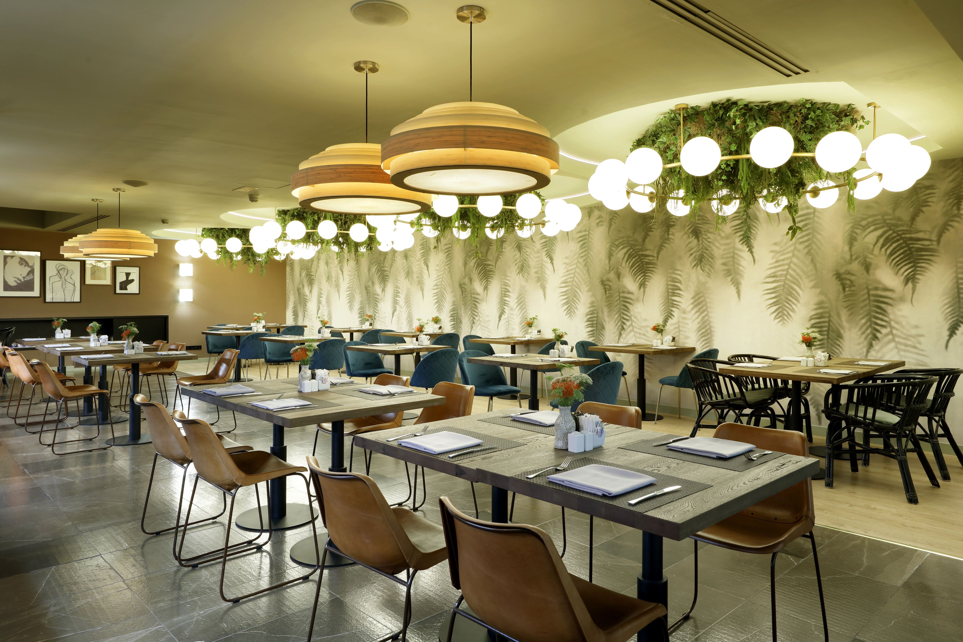 A spacious dining area with multiple tables and chairs, modern pendant lighting, and a decorative ceiling with hanging plants.
