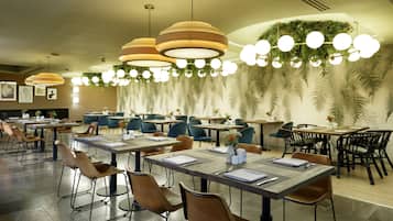 A spacious dining area with multiple tables and chairs, modern pendant lighting, and a decorative ceiling with hanging plants.