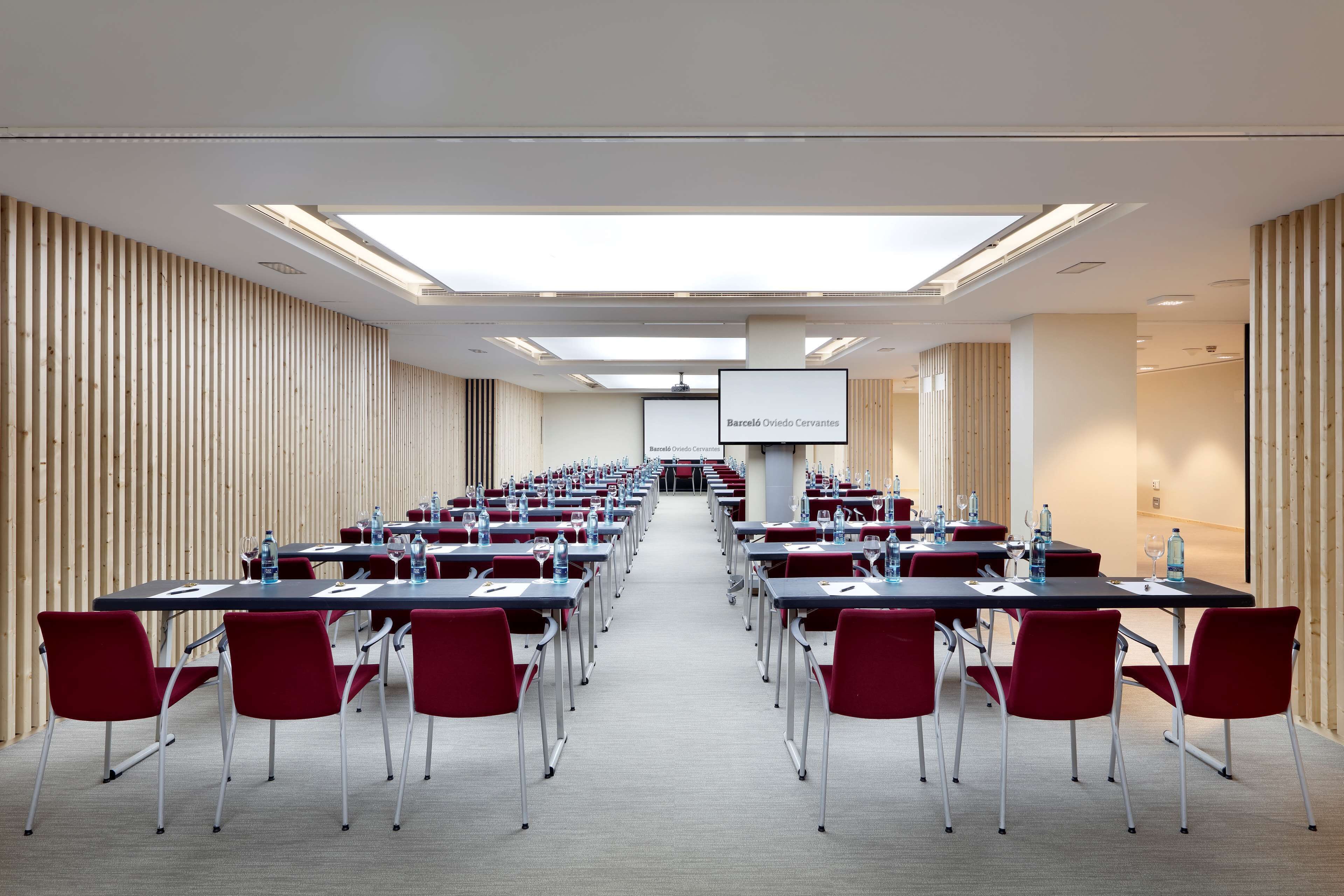 A conference room with rows of tables and chairs, a projector screen, and a wooden slat wall.