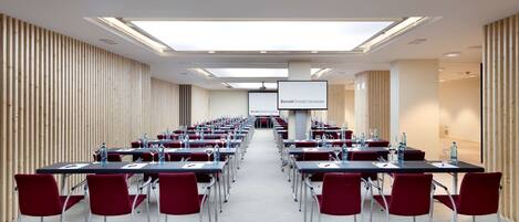 A conference room with rows of tables and chairs, a projector screen, and a wooden slat wall.