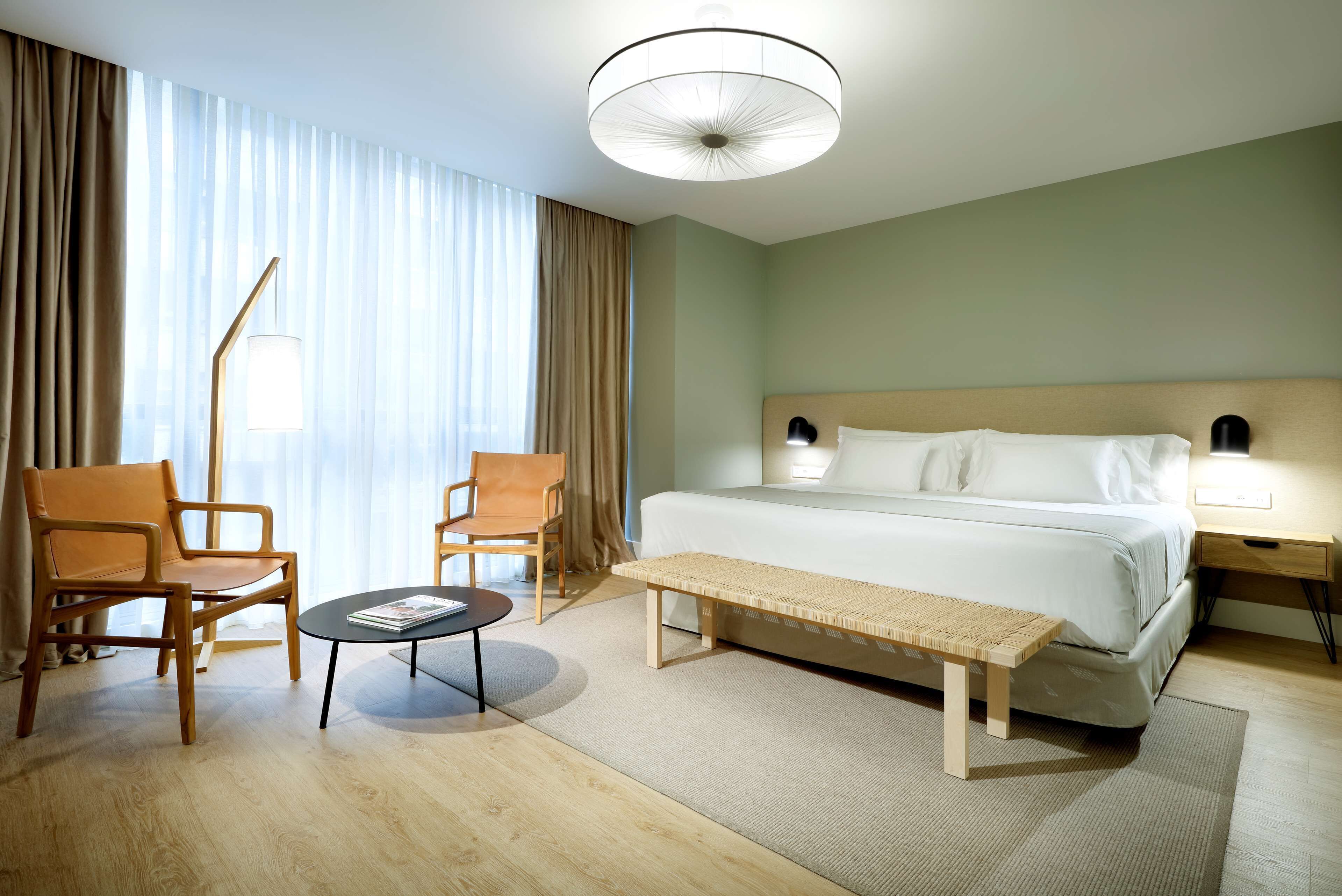 Quarto de hotel com cama, duas cadeiras, uma mesinha, um banco e uma mesa de cabeceira com abajur.