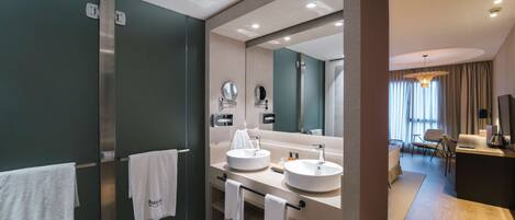 A modern bathroom with two sinks, a large mirror, and a vanity. There are towels hanging on a rack and a shower area visible.