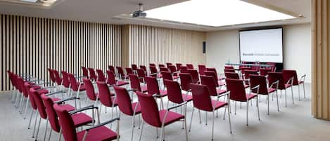 Een congreszaal met rijen rode stoelen, een scherm voor een beamer en een wand van houten latten.