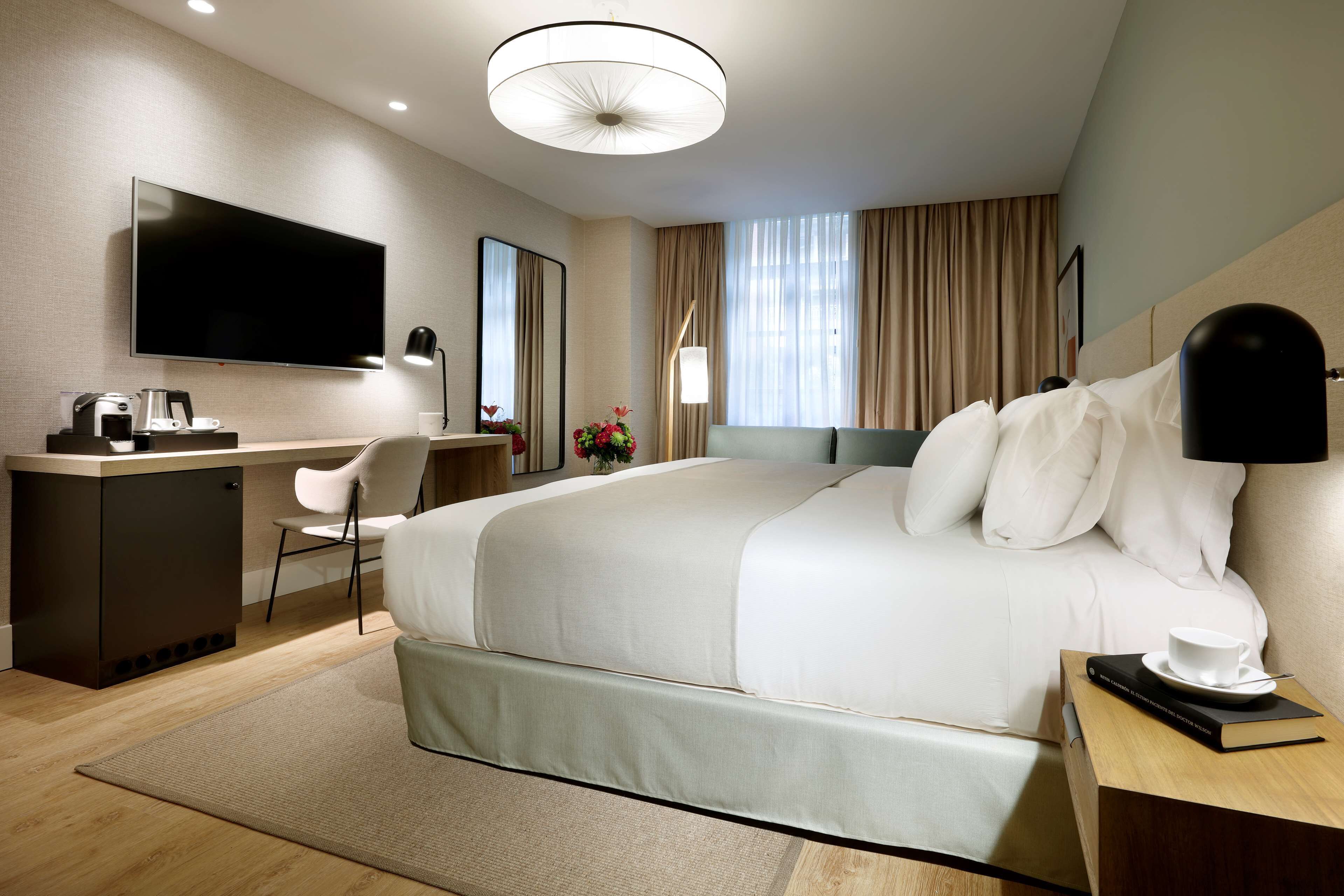 Una habitación de hotel moderna con una cama grande, un escritorio con televisor de pantalla plana, una cafetera y una mesita de noche con un libro y una taza.