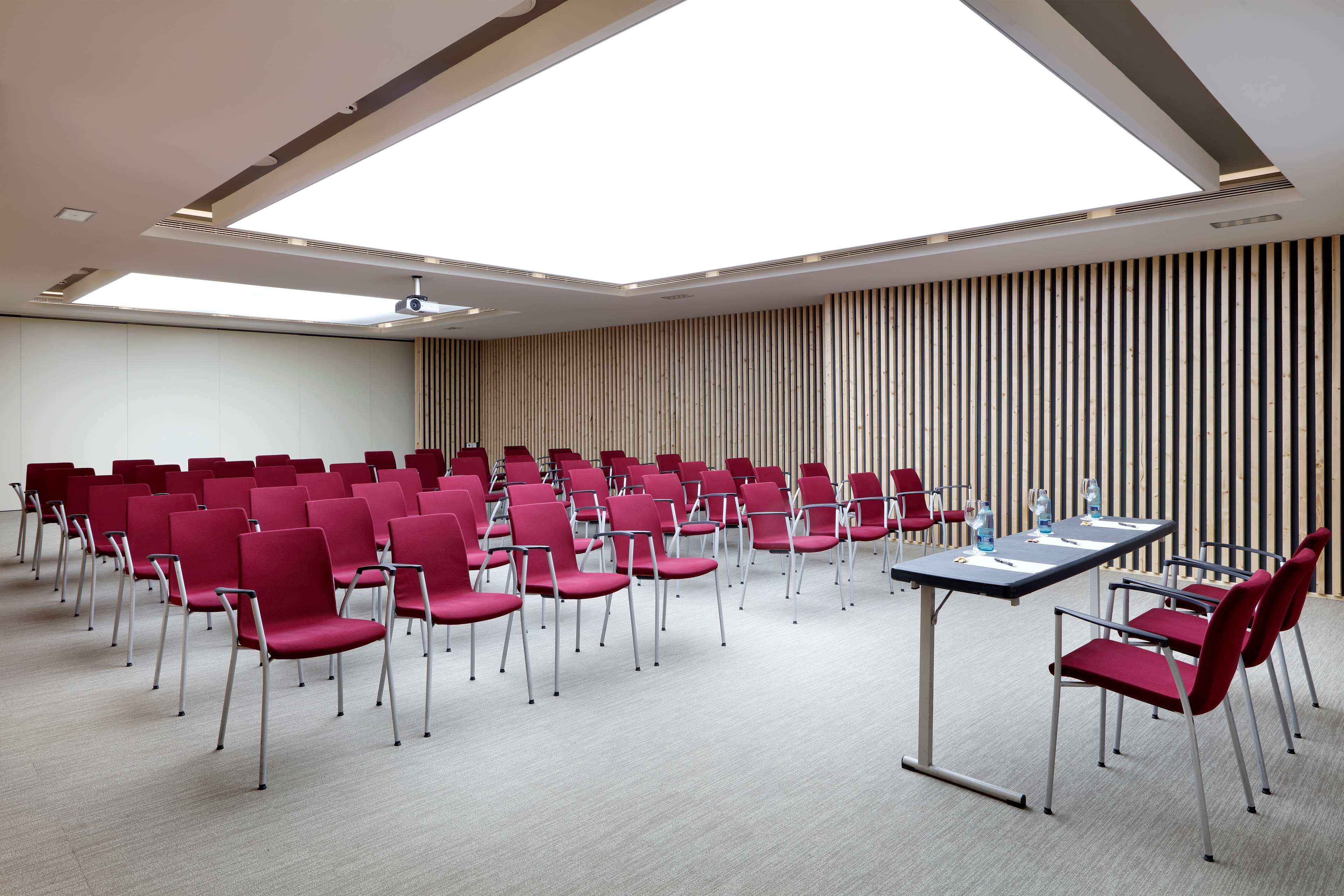 Un salón de conferencias con filas de sillas rojas, una mesa larga y un techo inclinado con tragaluces.