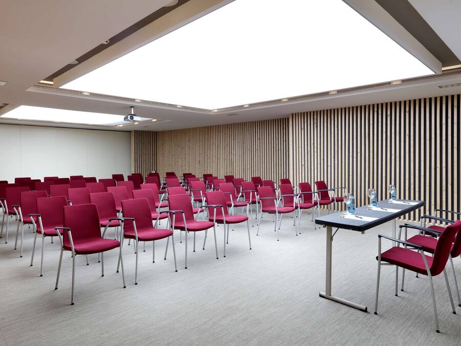 Un salón de conferencias con filas de sillas rojas, una mesa larga y un techo inclinado con tragaluces.
