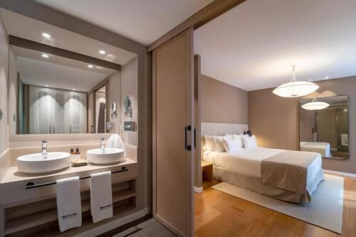 Une chambre d’hôtel moderne avec un grand lit, une salle de bain séparée dotée d’un lavabo et d’un grand miroir.