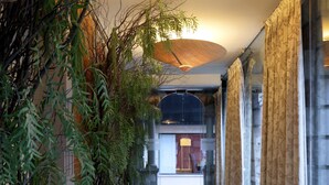 A corridor with a large woven ceiling light, potted plants, and a view of a room with a bed and a nightstand.