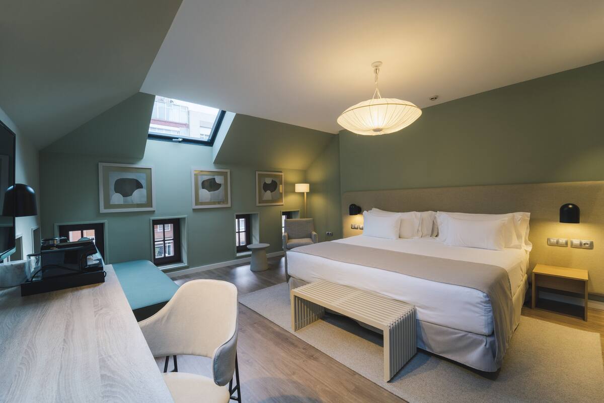 Een moderne slaapkamer met een groot bed, een bureau en een stoel. Er zijn wanddecoraties met ingelijste foto's, een dakraam en een hanglamp.