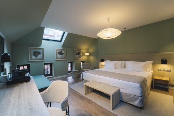 A modern bedroom with a large bed, a desk, and a chair. There are framed pictures on the wall, a skylight, and a hanging light fixture.