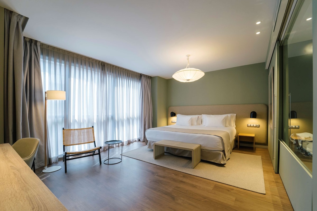 Une chambre d’hôtel avec un lit, des lampes de chevet, une chaise, une petite table et une grande fenêtre avec des rideaux.