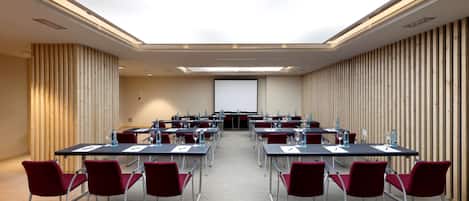 Un salón de conferencias con una larga mesa, sillas rojas y una pantalla de proyector al frente.