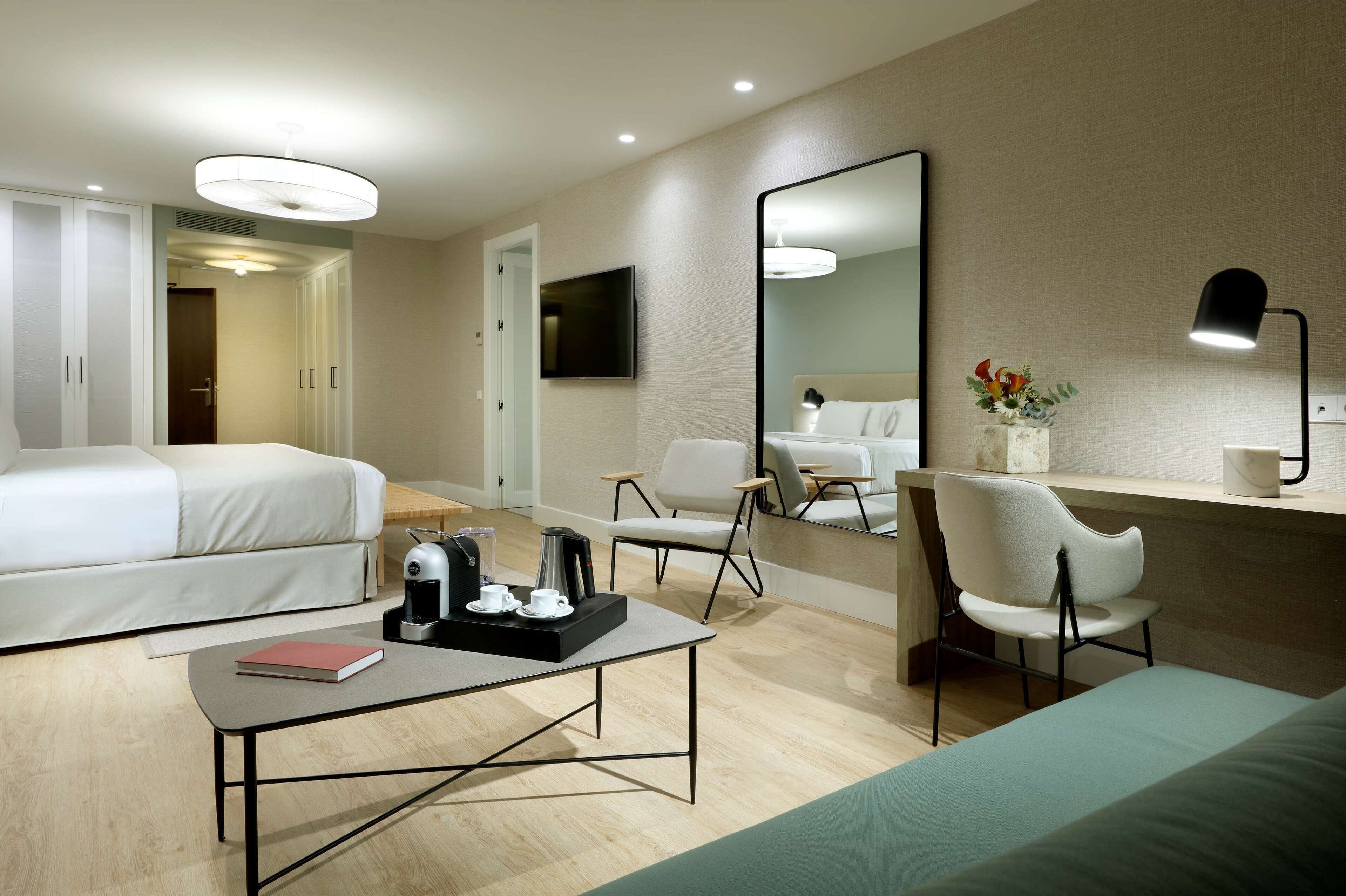 A hotel room with a bed, a desk with a coffee maker, a chair, a TV, and a large mirror.