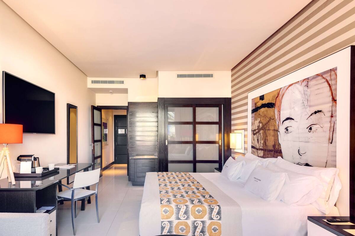 Une chambre d’hôtel moderne dotée d’un grand lit, d’un bureau avec une chaise, d’un téléviseur à écran plat et d’un grand tableau abstrait accroché au mur.