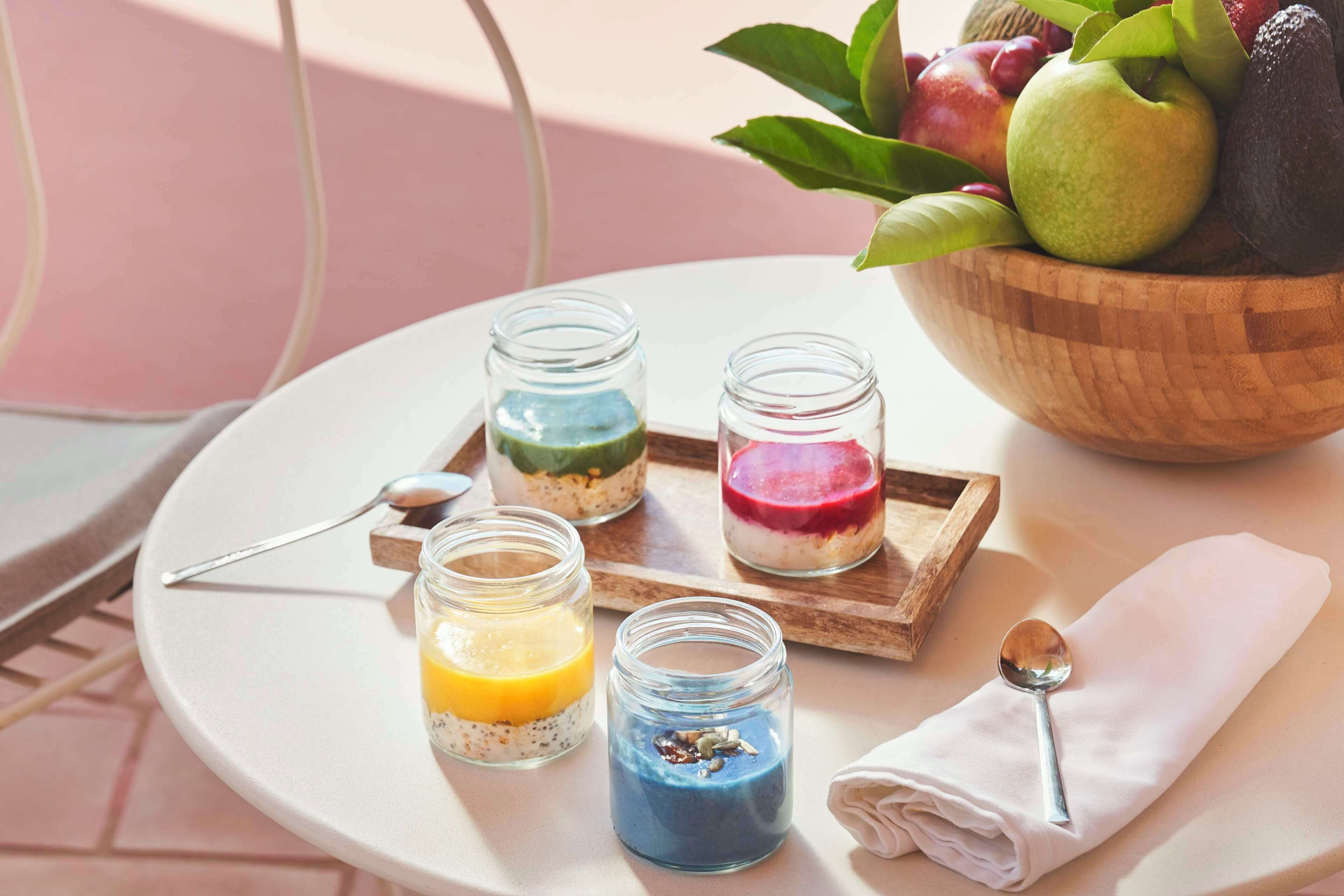 Ein Tisch mit vier Gläsern, die farbenfrohe, geschichtete Desserts enthalten, einer Holzschüssel mit Früchten und einem Löffel auf einer Serviette.