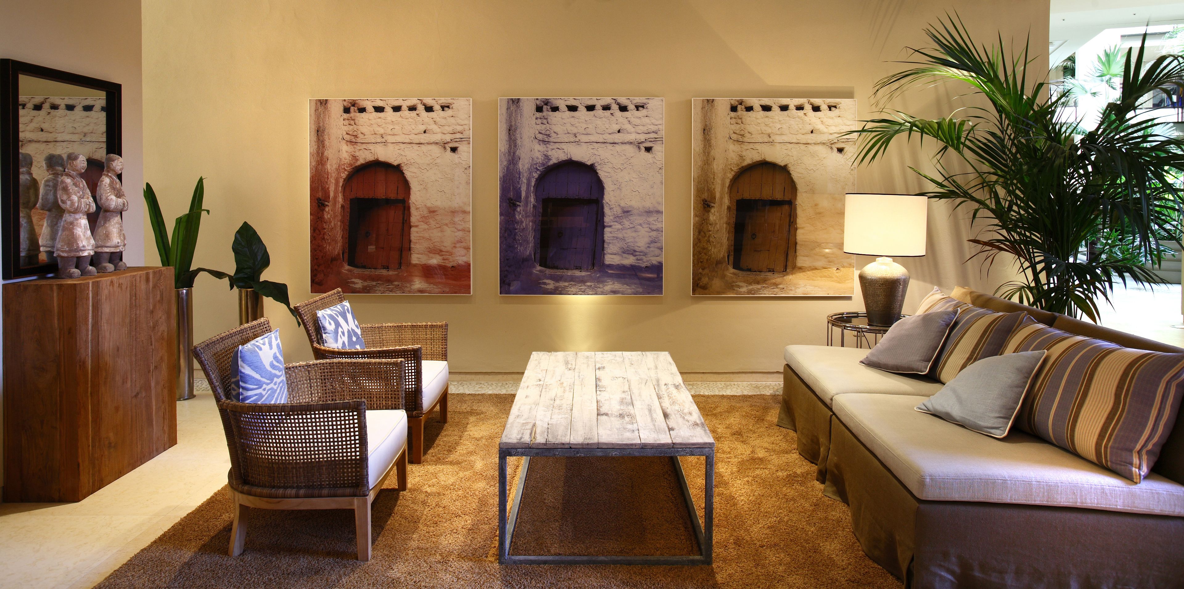 A cozy living room with wicker furniture, a wooden coffee table, and a sofa with striped cushions. There are three framed artworks depicting arched doorways on the wall.
