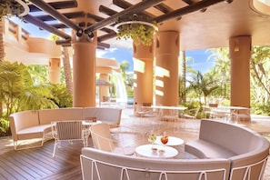 A covered outdoor seating area with tables and chairs, surrounded by palm trees and a waterfall view.
