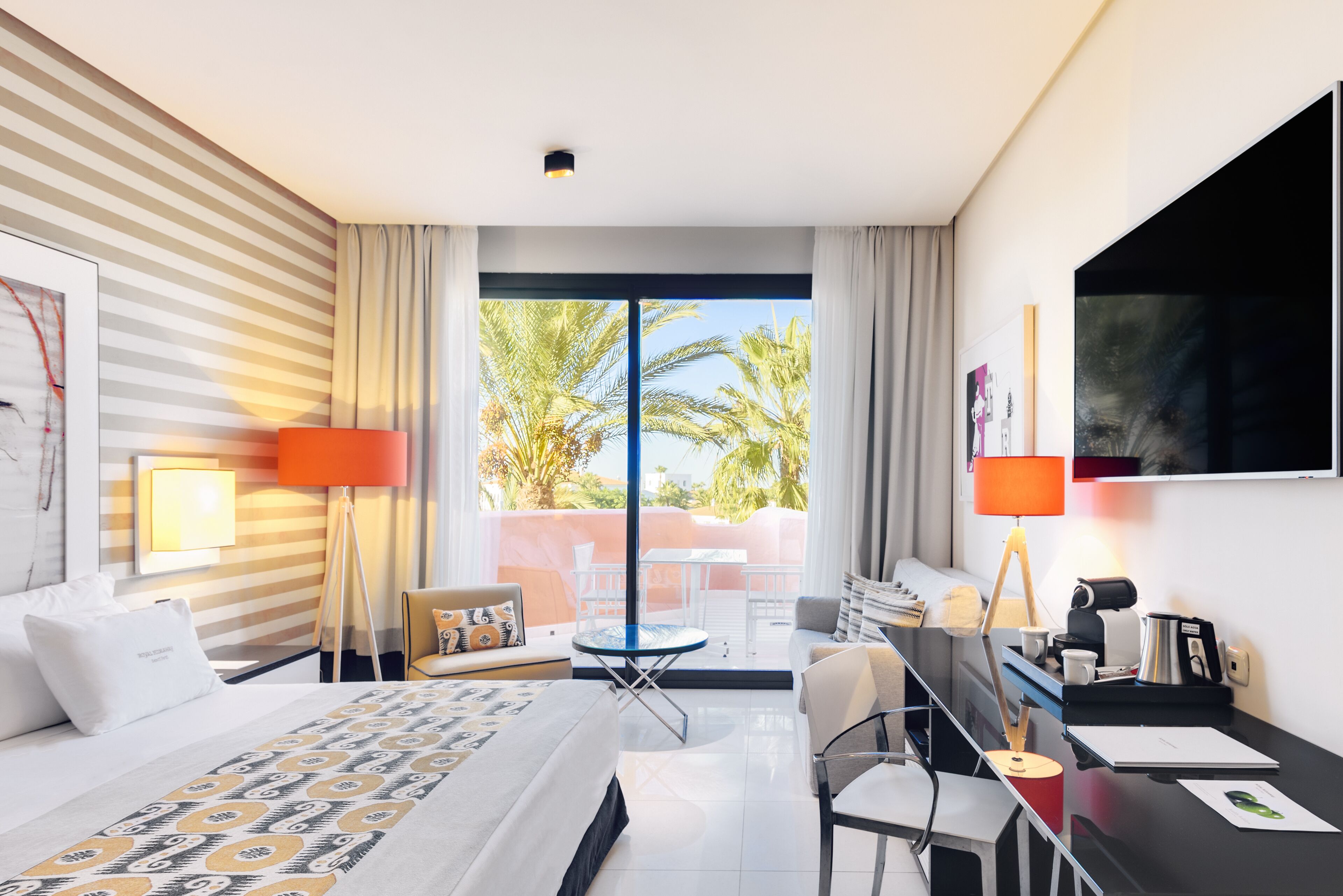 Habitación de hotel con cama, escritorio, silla, televisor y balcón con vista a palmeras.