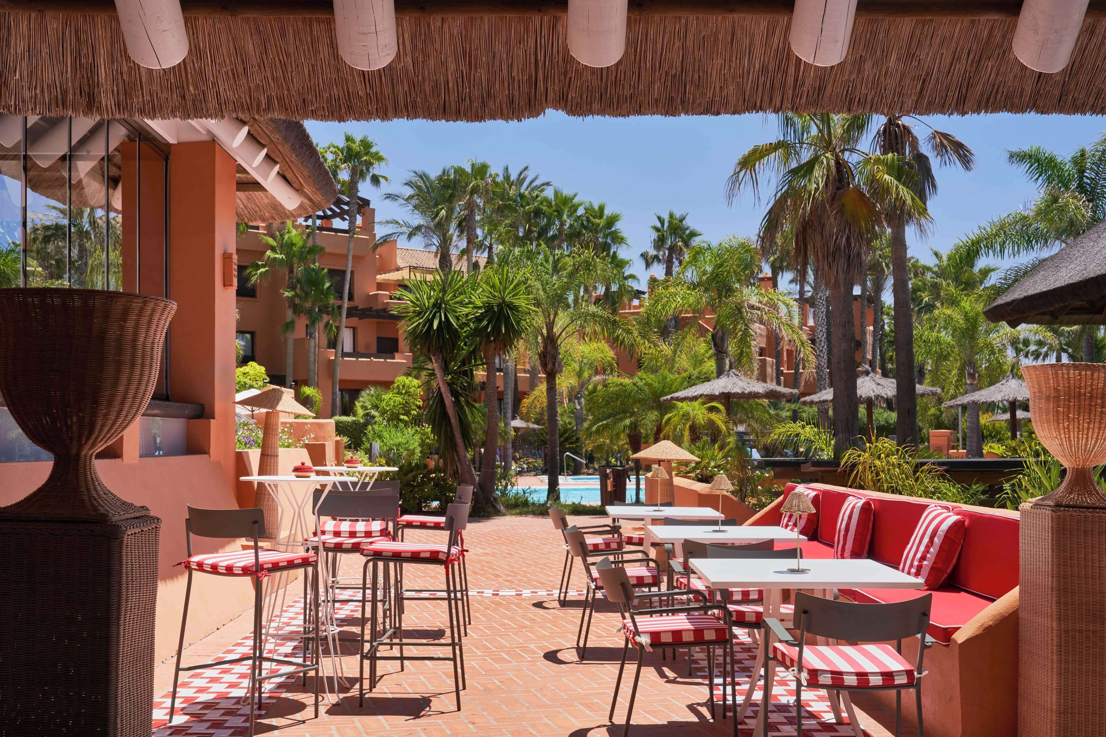 Un patio al aire libre con mesas y sillas, cojines a cuadros rojos y blancos, y sombrillas de paja.