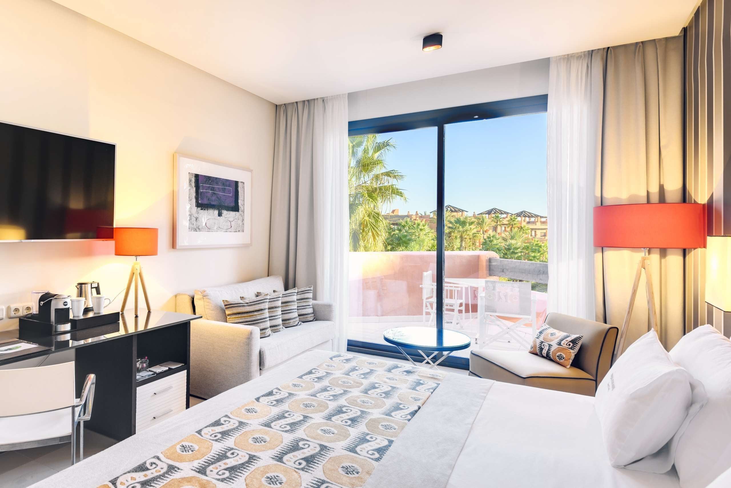 Une chambre d’hôtel moderne dotée d’un grand lit, d’un bureau, d’une chaise, d’un canapé et d’un balcon offrant une vue sur les palmiers et les immeubles.