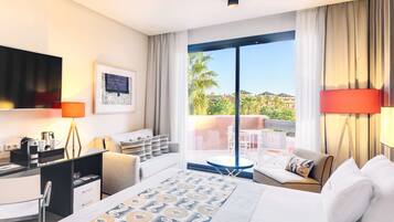 A modern hotel room with a large bed, a desk, a chair, a sofa, and a balcony with a view of palm trees and buildings.