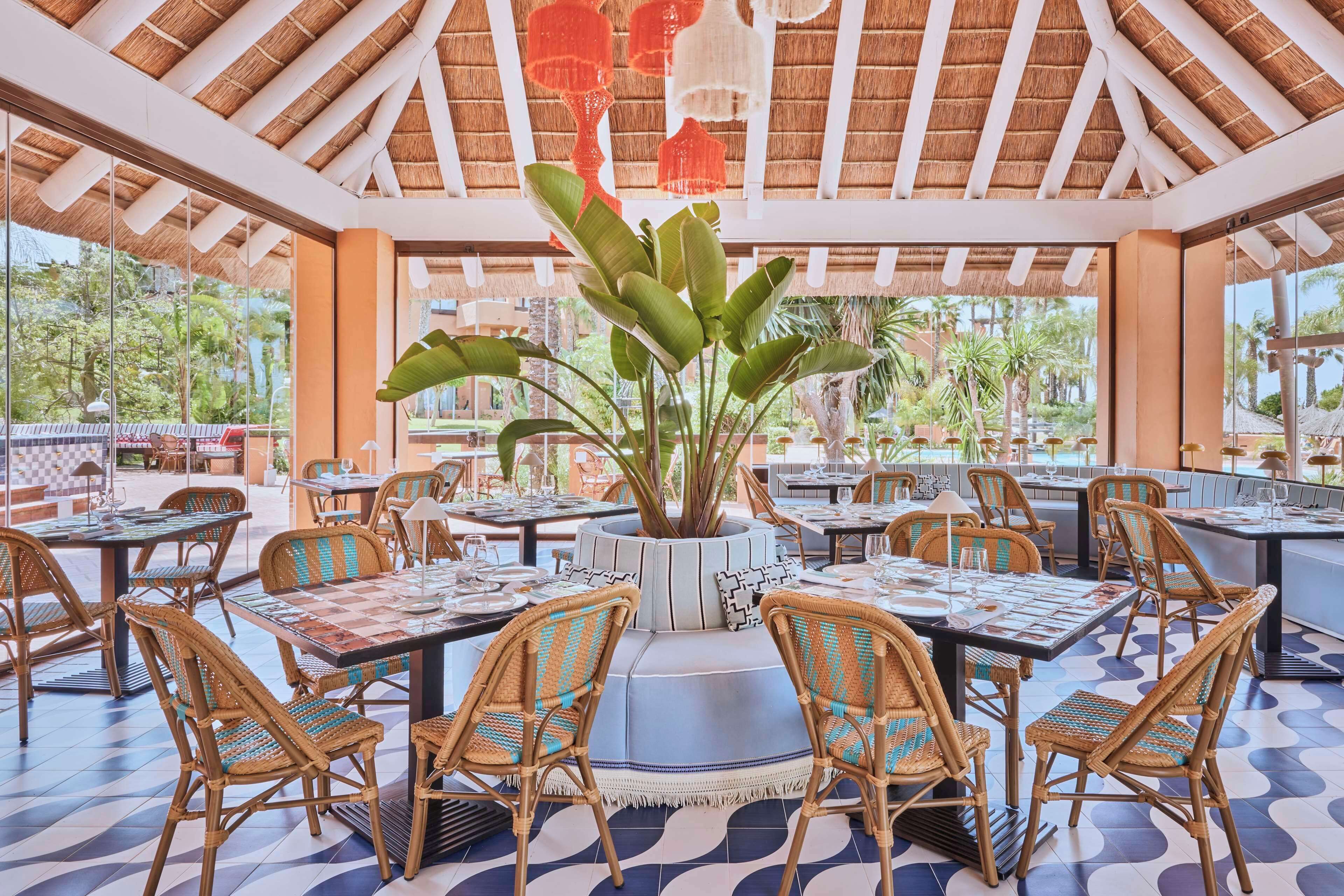 An indoor dining area with tables and chairs, a central plant, and a view of the outdoors.