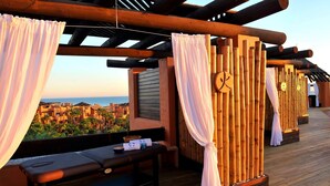 Eine Holzterrasse mit einer Massageliege, weißen Vorhängen und einer Bambuswand.