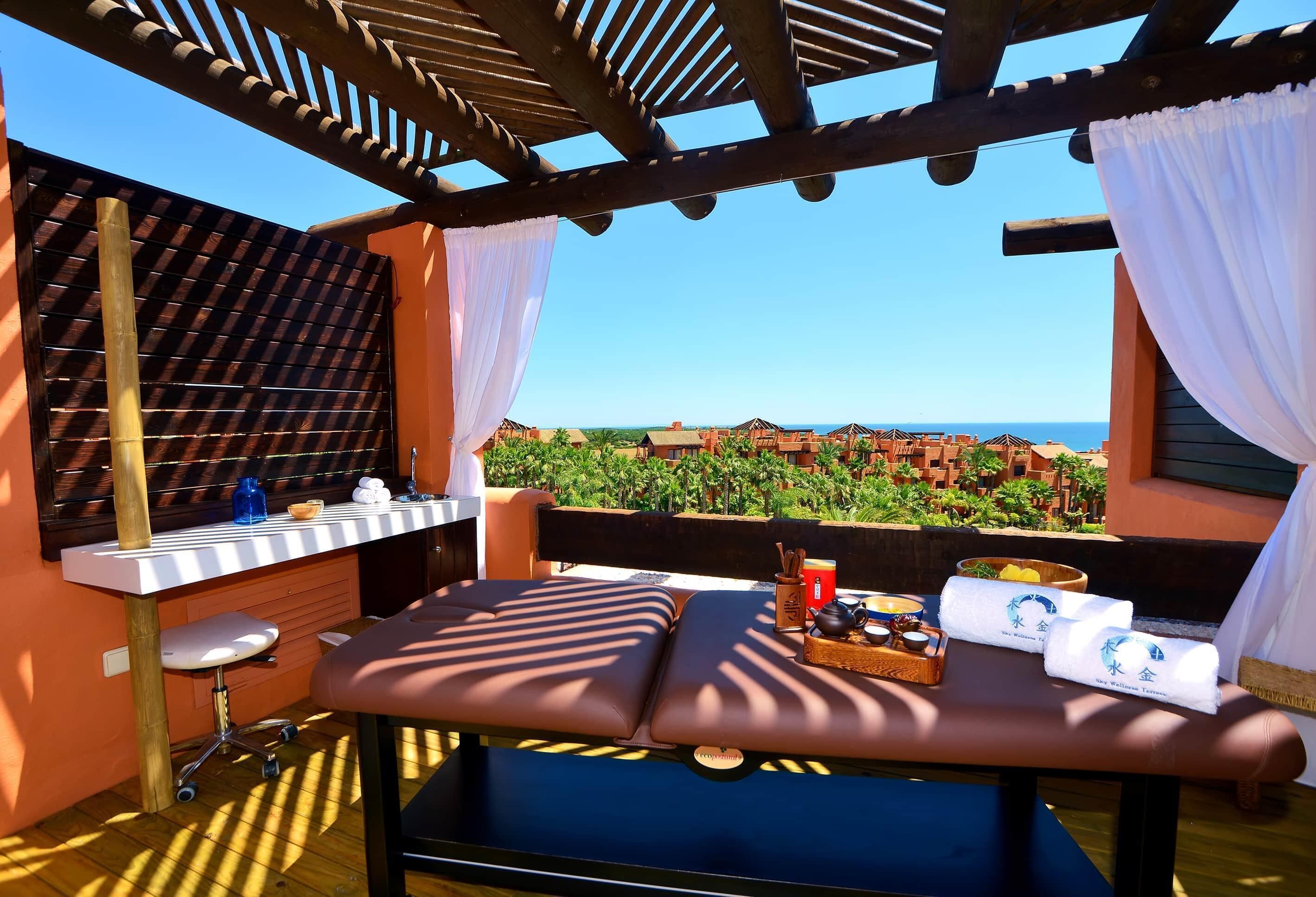 A sunlit outdoor seating area with a massage table, a small desk, and a view of a vineyard and ocean.