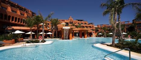 A large outdoor swimming pool surrounded by palm trees and a hotel with multiple balconies.
