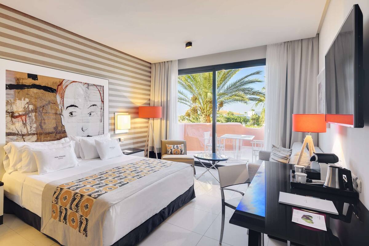 Une chambre d’hôtel avec un grand lit, un bureau, une chaise, une télévision et un balcon offrant une vue sur les palmiers.