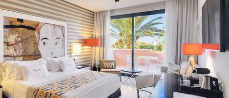 A hotel room with a large bed, a desk, a chair, a TV, and a balcony with a view of palm trees.